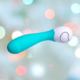 The OhMiBod Lovelife Cuddle Mini G-Spot Vibrator in turquoise, featuring a rounded handle and control buttons, is set against a blurred background of colorful lights, making it the perfect travel companion.